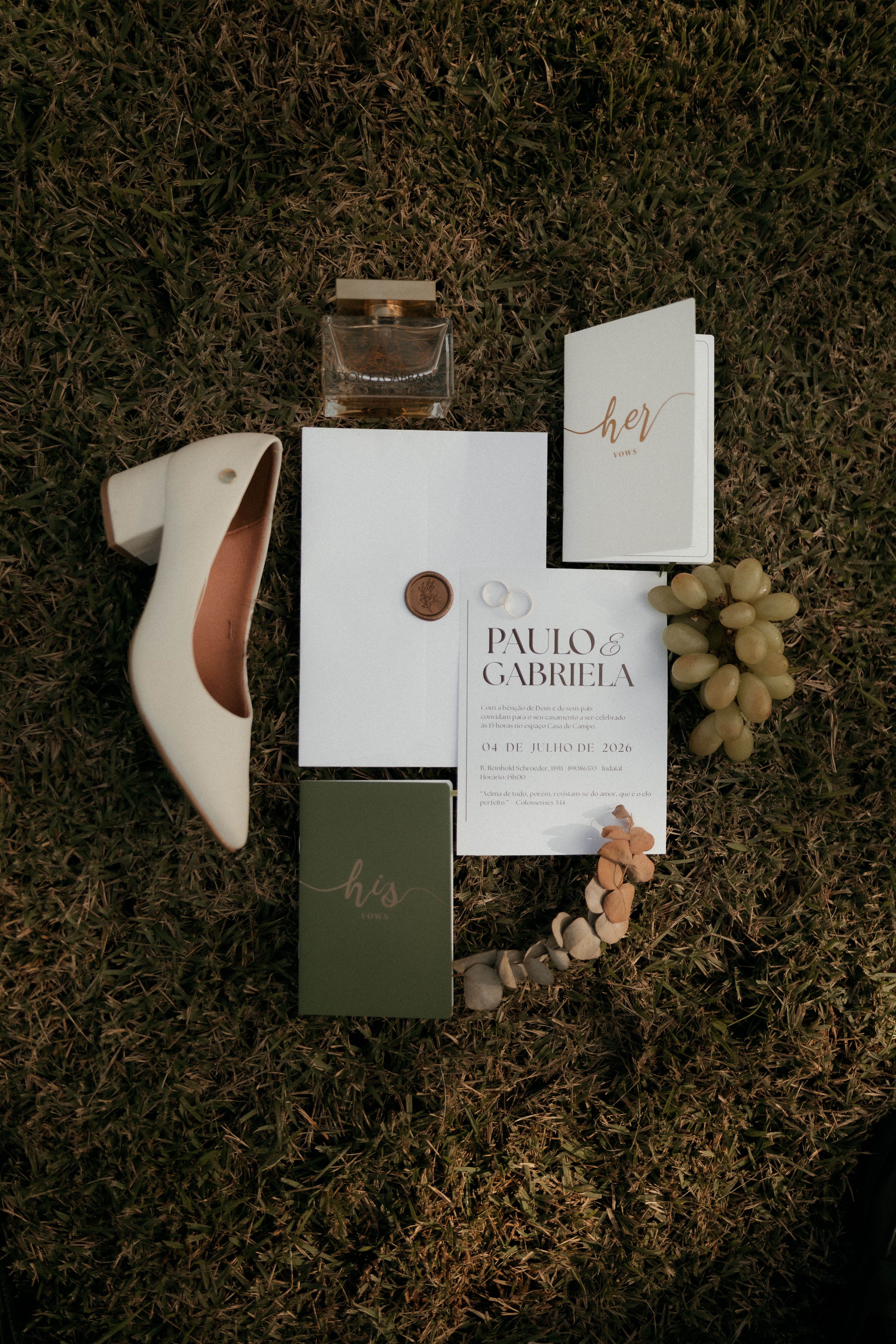 Flat lay of wedding invitation with shoes, perfume, and decor elements on grass in natural light.