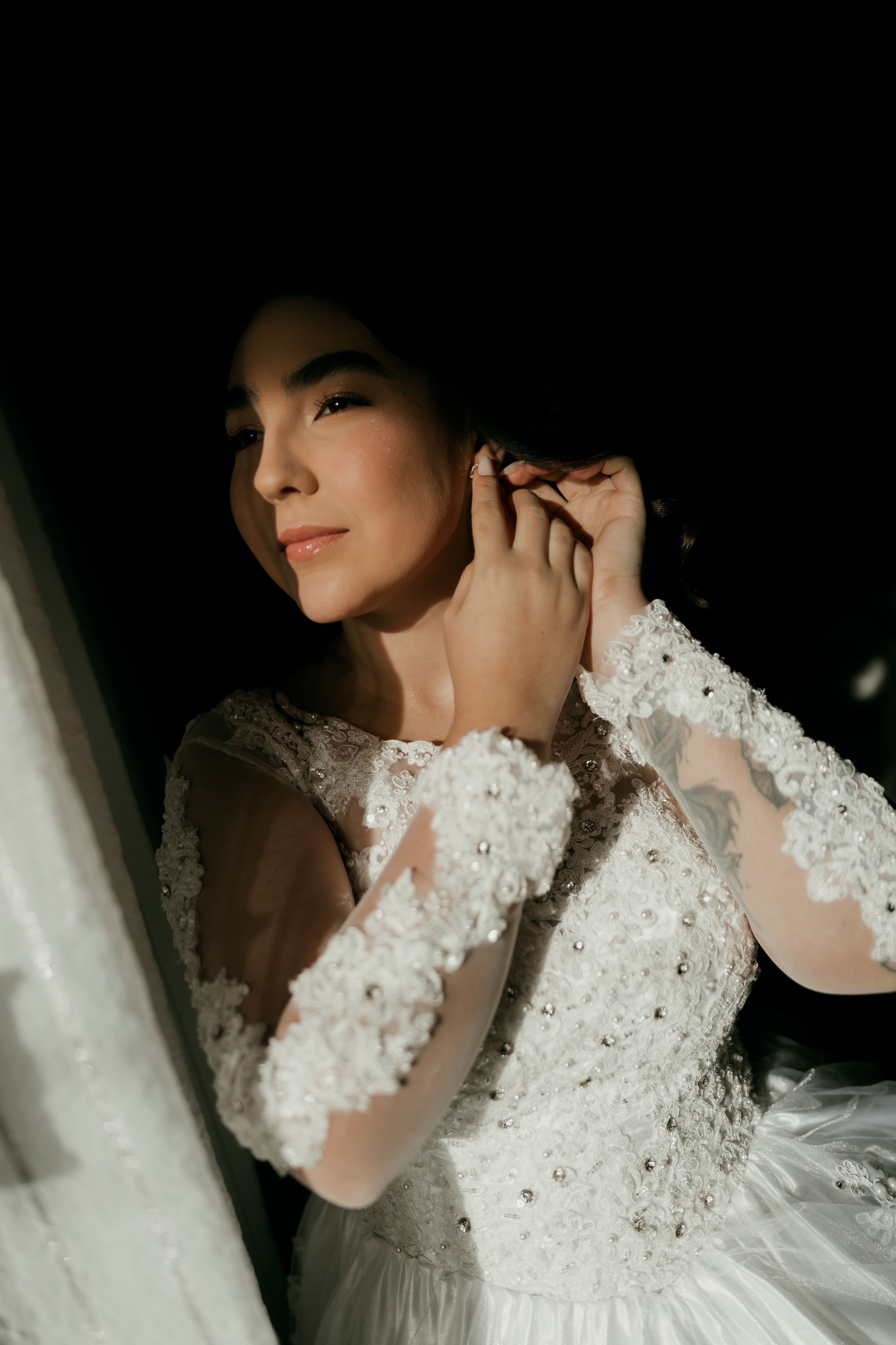 Elegant bride in white lace gown adjusts earrings, bathed in soft light, portraying a serene pre-wedding moment.