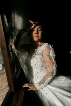 A bride in a lace dress poses by a sunlit window, showcasing artistic shadow play.