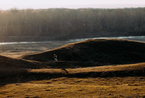 Free A person runs across hilly terrain at sunrise beside a tranquil river. Stock Photo
