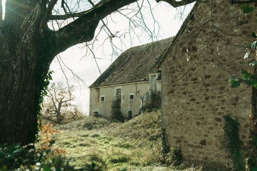 Abandoned stone building in winter, La Berthenoux, France, surrounded by bare trees and overgrown grass.