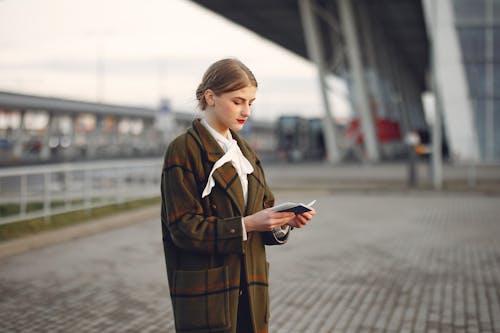 Attentive female passenger wearing trendy plaid coat and white blouse checking passport and ticket standing on pavement near modern building of airport outside