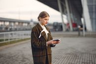 Attentive female passenger wearing trendy plaid coat and white blouse checking passport and ticket standing on pavement near modern building of airport outside
