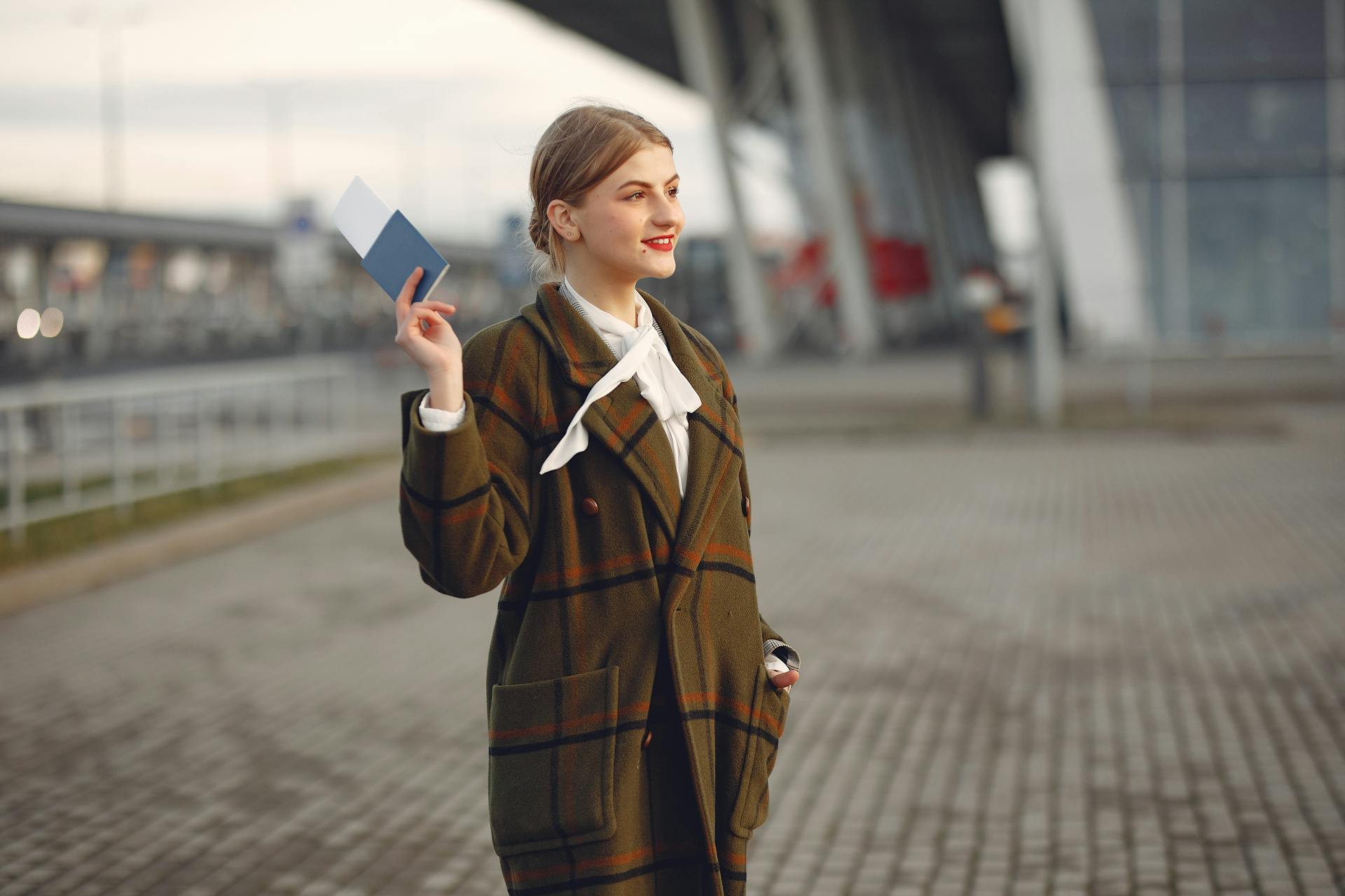 Cheerful female passenger wearing trendy plaid coat taking passport and ticket in raised hand while standing on pavement near modern building of airport outside and looking away with smile