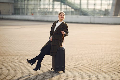 Free Cheerful stylish woman sitting on suitcase in front of contemporary glass building on urban street Stock Photo