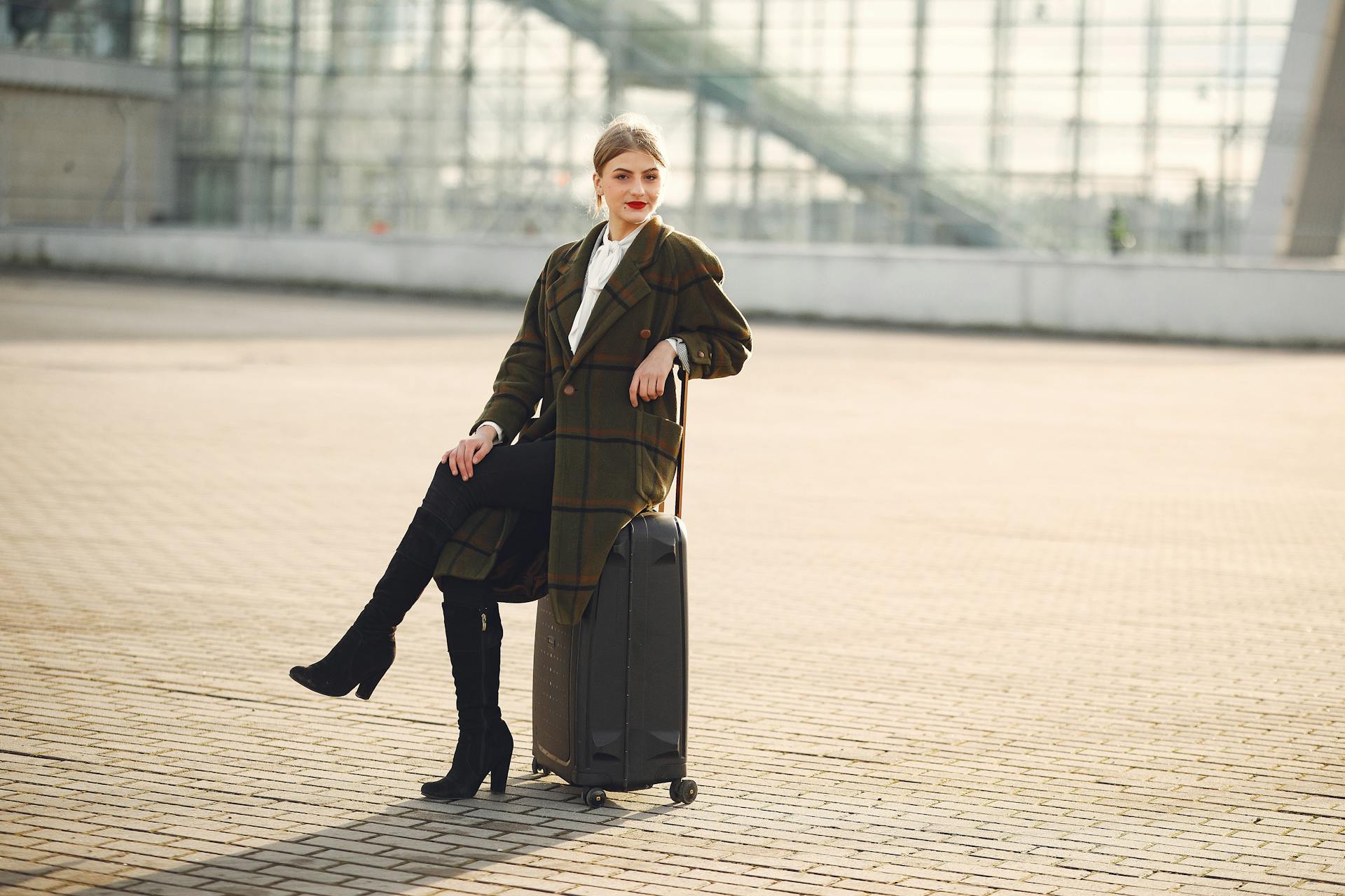 Full length of positive female traveler wearing warm trendy plaid coat and high boots sitting on suitcase with legs crossed on pavement near modern airport terminal and smiling looking at camera in daylight during cold season