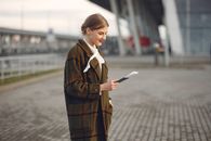 Smiling female passenger wearing trendy plaid coat and white blouse checking passport and ticket standing on pavement near contemporary building of airport