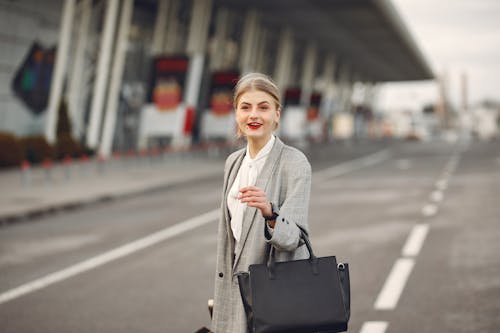Positive female passenger wearing formal plaid suit standing on asphalt road to airport with handbag and suitcase while smiling looking at camera during business trip