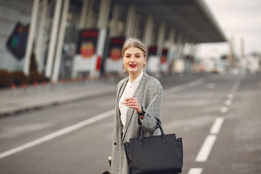 Positive female passenger wearing formal plaid suit standing on asphalt road to airport with handbag and suitcase while smiling looking at camera during business trip