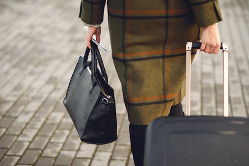 Free Back view of unrecognizable young female traveler in warm trendy plaid coat strolling on pavement with suitcase and bag while taking passport and tickets on city street in cold season Stock Photo