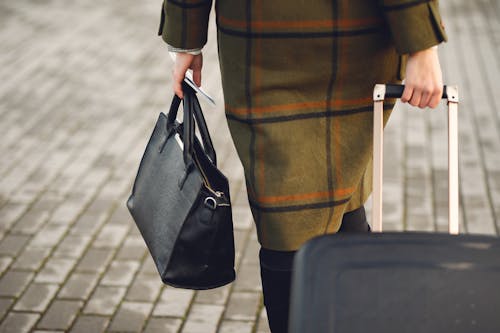 Back view of unrecognizable young female traveler in warm trendy plaid coat strolling on pavement with suitcase and bag while taking passport and tickets on city street in cold season