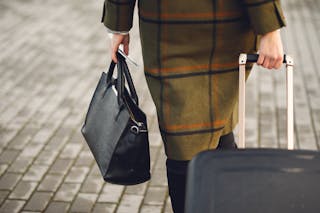 Back view of unrecognizable young female traveler in warm trendy plaid coat strolling on pavement with suitcase and bag while taking passport and tickets on city street in cold season