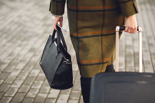 Free Back view of unrecognizable young female traveler in warm trendy plaid coat strolling on pavement with suitcase and bag while taking passport and tickets on city street in cold season Stock Photo
