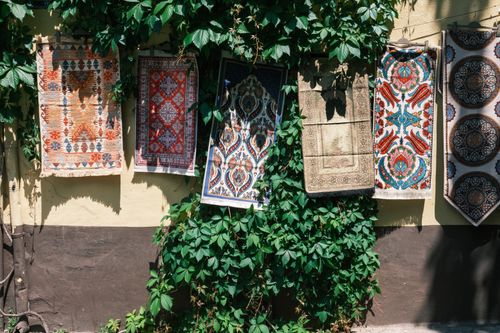Free Colorful tapestries with intricate designs hanging on a wall amidst green vines, capturing a vibrant summer day. Stock Photo