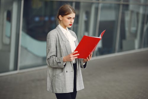 Young female entrepreneur in stylish formal wear reading documents while standing near modern business building in city street