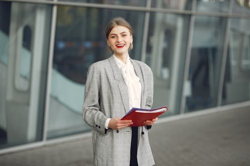 Free Positive young woman in trendy wear smiling at camera while holding documents standing against glass building in downtown Stock Photo