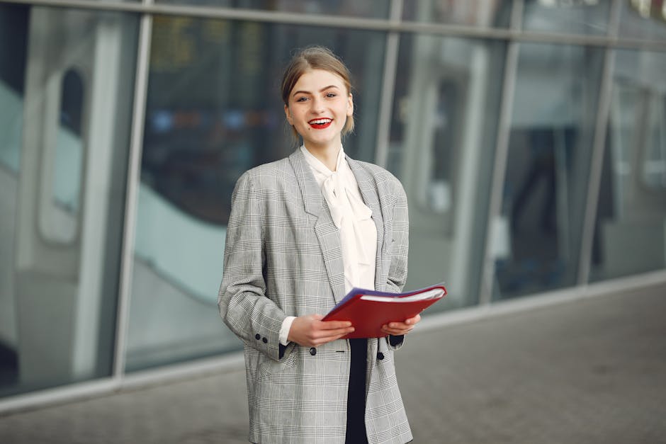 Positive young woman in trendy wear smiling at camera while holding documents standing against glass building in downtown