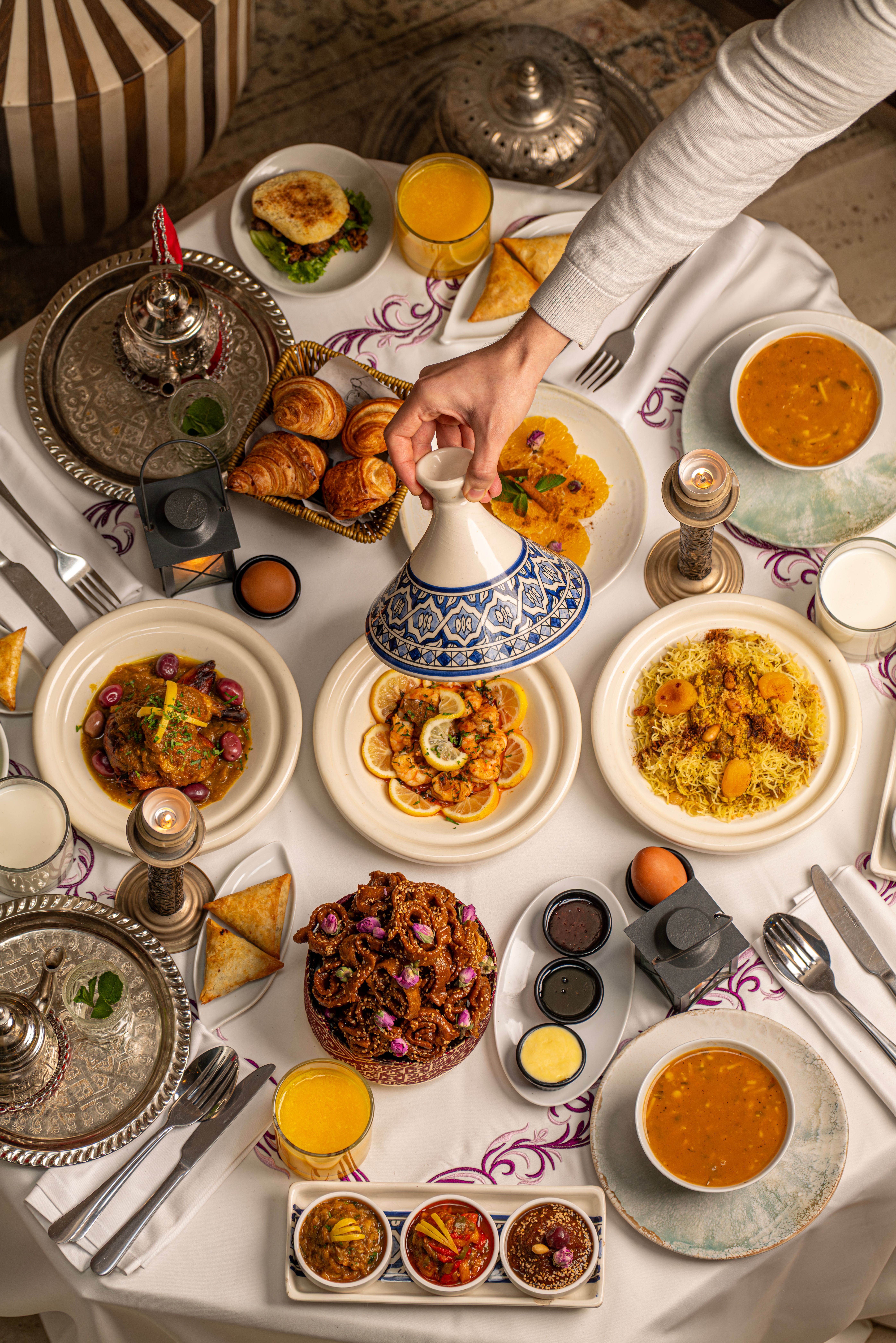 Table vue de dessus : une main soulève le couvercle bleu et blanc d’un tajine, poulet aux olives et au citron, crevettes au citron, plat de vermicelles aux amandes et aux abricots, pâtisseries au miel