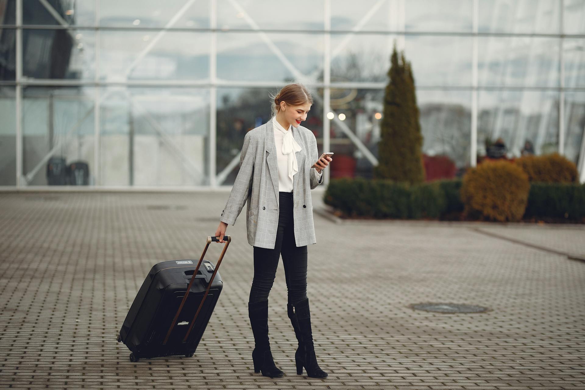 Confident businesswoman using smartphone while traveling with luggage outside an airport terminal.