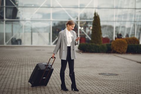 Full length of stylish woman in elegant outfit carrying baggage and surfing smartphone while standing near glass wall of airport