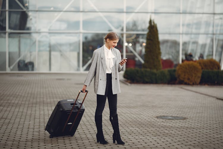 Smiling Female Passenger With Suitcase Messaging On Mobile Phone In Street