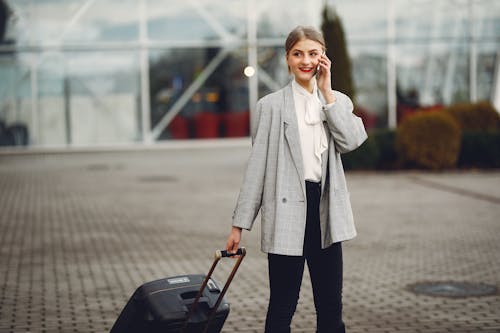 Stylish businesswoman speaking on smartphone while standing with luggage near airport