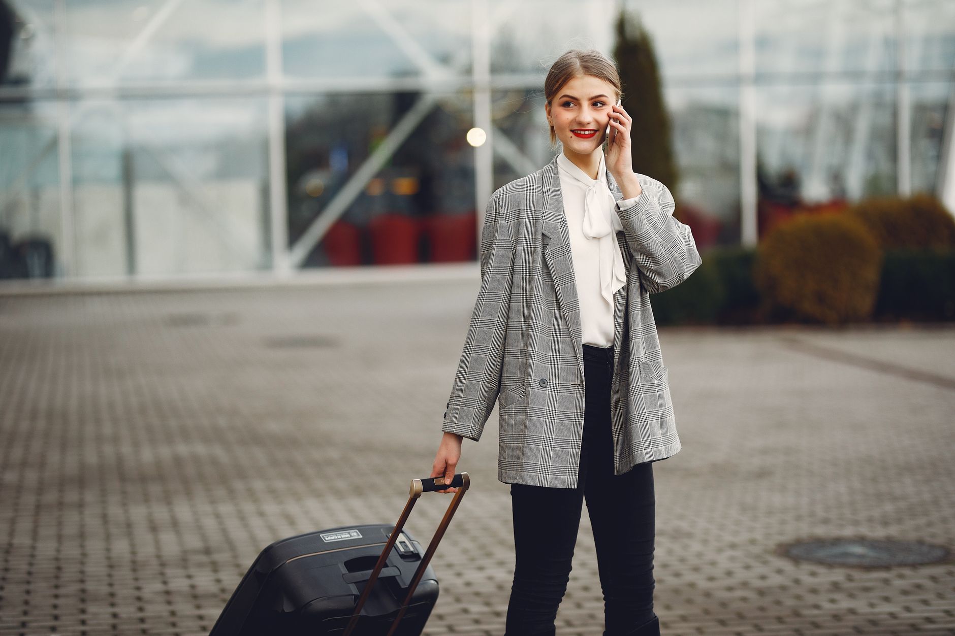 Positive female in trendy outfit talking on phone and looking away while waiting for taxi with suitcase near airport terminal