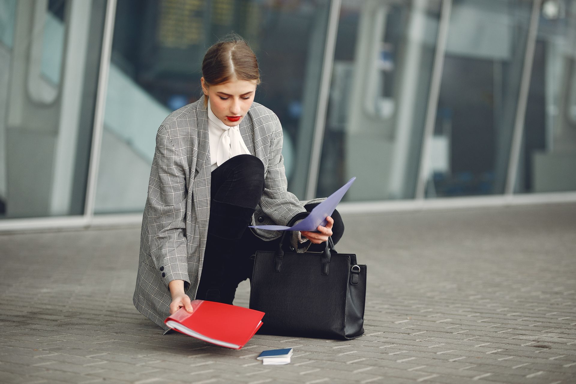 Distracted female manager in trendy wear collecting scattered documents and passport from sidewalk into briefcase near glass wall of modern building