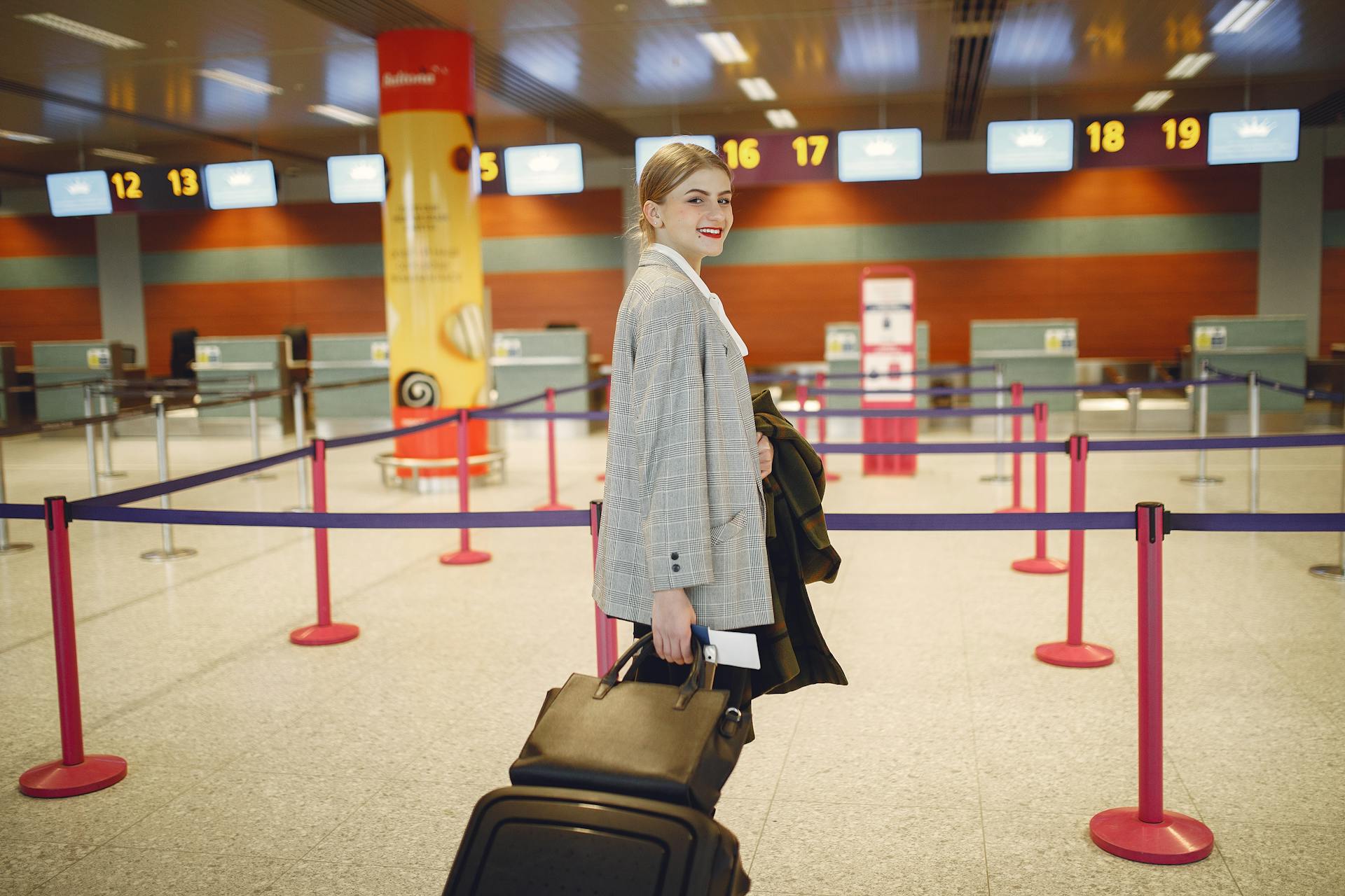 Side view of happy businesswoman in trendy jacket carrying baggage and smiling at camera while waiting for flight in modern airport