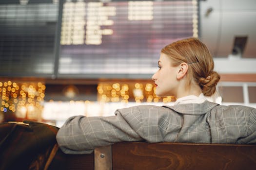 Pensive female designer in checkered jacket waiting for flight near departure board looking away in modern airport in evening