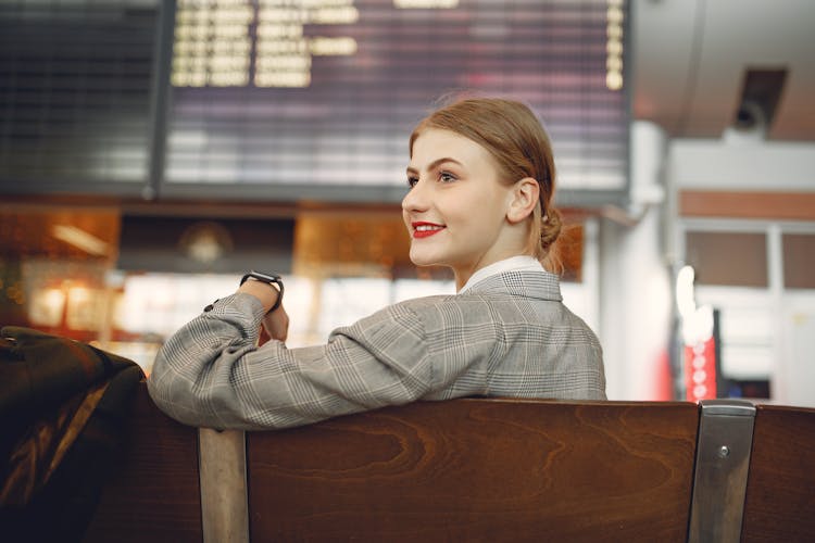 Side View Of Positive Female Manager Waiting For Flight On Wooden Seat In Airport