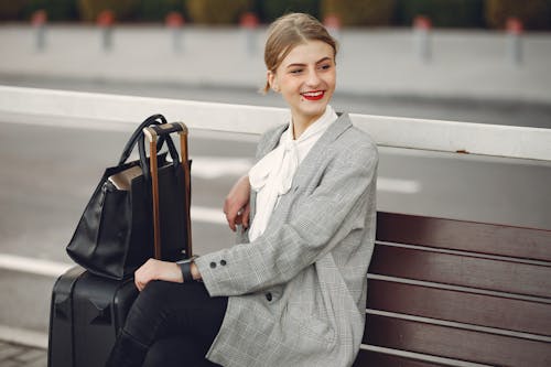 Positive female traveler in trendy clothes smiling away while waiting for train sitting on wooden bench with suitcase and bag in street