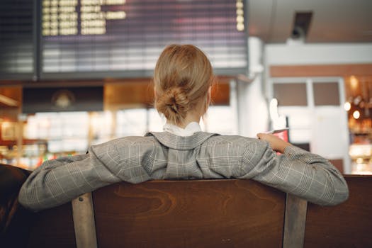 Back view of female employee in trendy jacket waiting for train departure and leaning on backs of wooden seats in train station