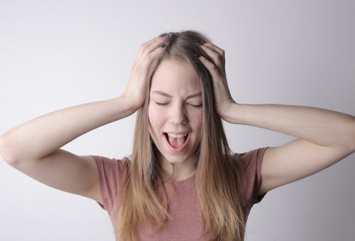 Angry crazy female in casual pink t shirt touching head while being unhappy and shouting loudly against gray wall background