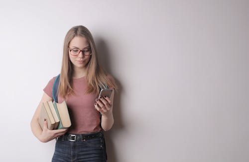 Free Confident intelligent female student in eyeglasses wearing pink t shirt and black jeans using cellphone while taking books standing against gray wall Stock Photo