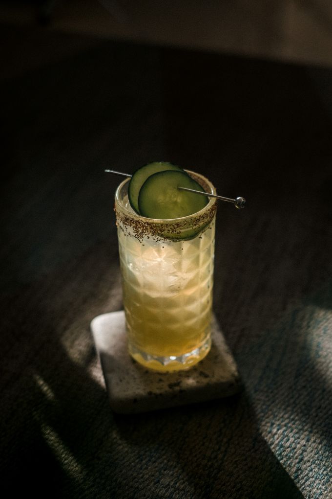 A cucumber-garnished cocktail on a stone coaster in low light, photo by emmanuel Lozano on Pexels