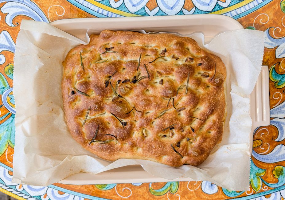 A golden focaccia loaf in a baking pan, its dimpled surface pooled with olive oil and scattered with rosemary sprigs and flaky salt, one corner cut into squares.