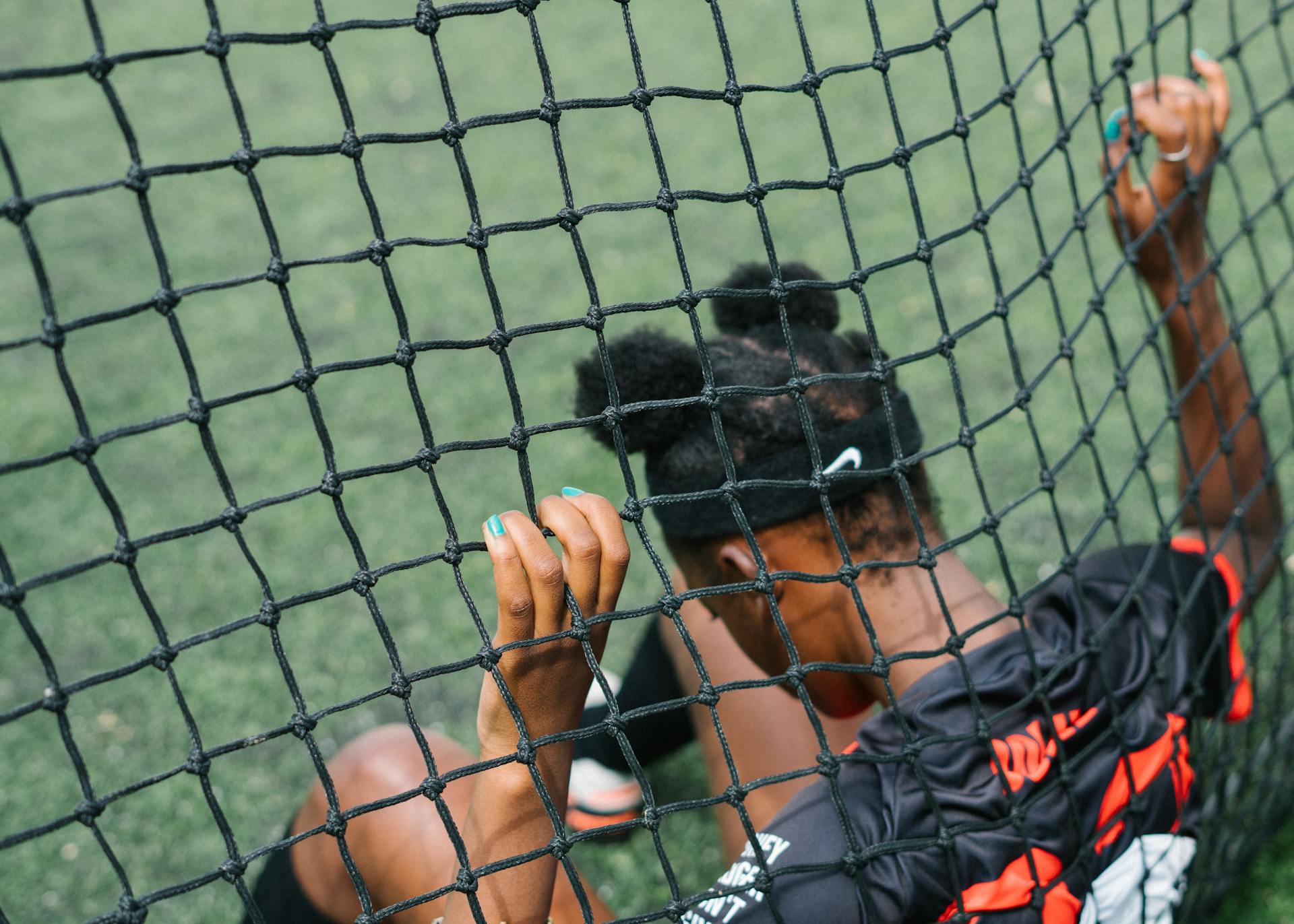 Back view of African American teenage football player sitting on field ground and holding grid of gate while preparing to start play football match