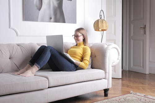 Woman In Yellow Long Sleeve Shirt And Blue Denim Jeans Sitting On White Sofa