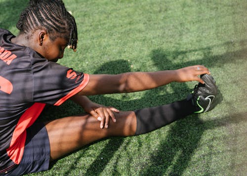 From above of black female soccer player sitting on green grass and stretching before coming on field