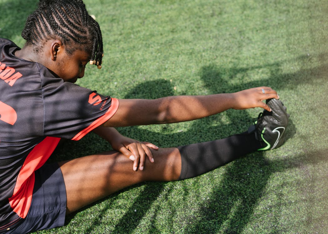 Free From above of black female soccer player sitting on green grass and stretching before coming on field Stock Photo