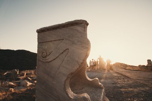 Marble sculpture in an ancient site at sunset with a group of tourists in the background.