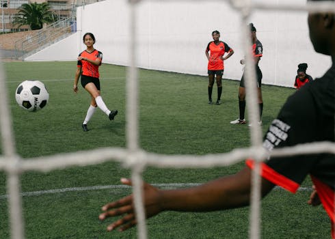 A group of female soccer players practice on a grassy field, one kicking the ball to the goalie.