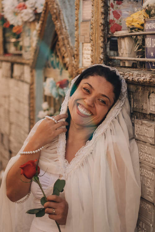 Free A smiling bride in white with a veil holds a red rose, creating a touching juxtaposition against a cemetery backdrop. Stock Photo