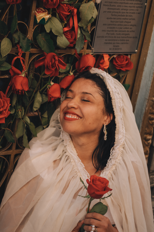 Free A bride in traditional attire joyfully holding a red rose, surrounded by flowers, exuding happiness. Stock Photo