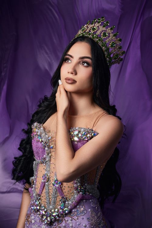 Free A woman in an ornate violet gown and crown gazes confidently, set against a purple backdrop. Stock Photo
