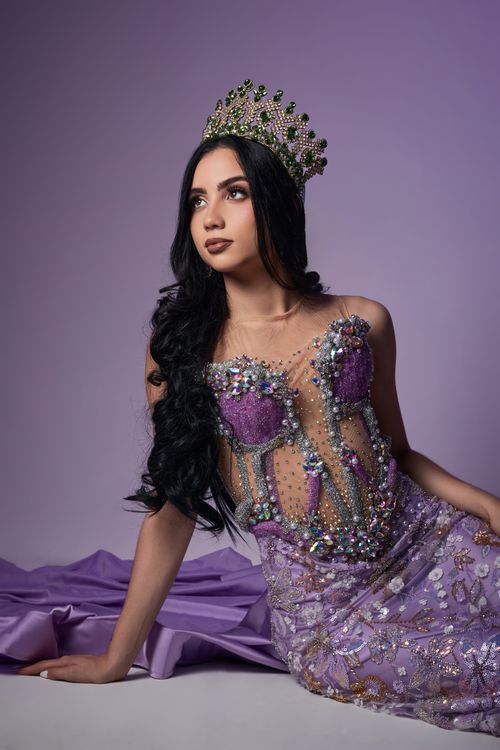 Free Beautiful woman in a purple dress and ornate crown posing gracefully against a purple backdrop. Stock Photo