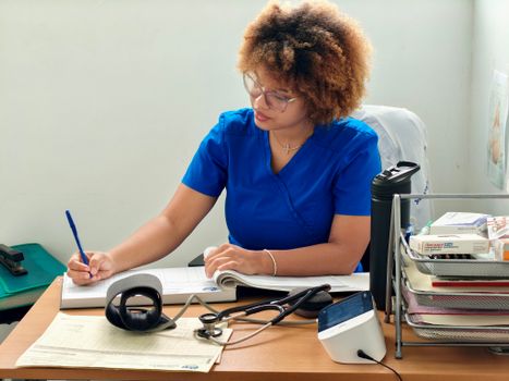 A nurse in blue scrubs writing patient information at a desk with medical equipment.