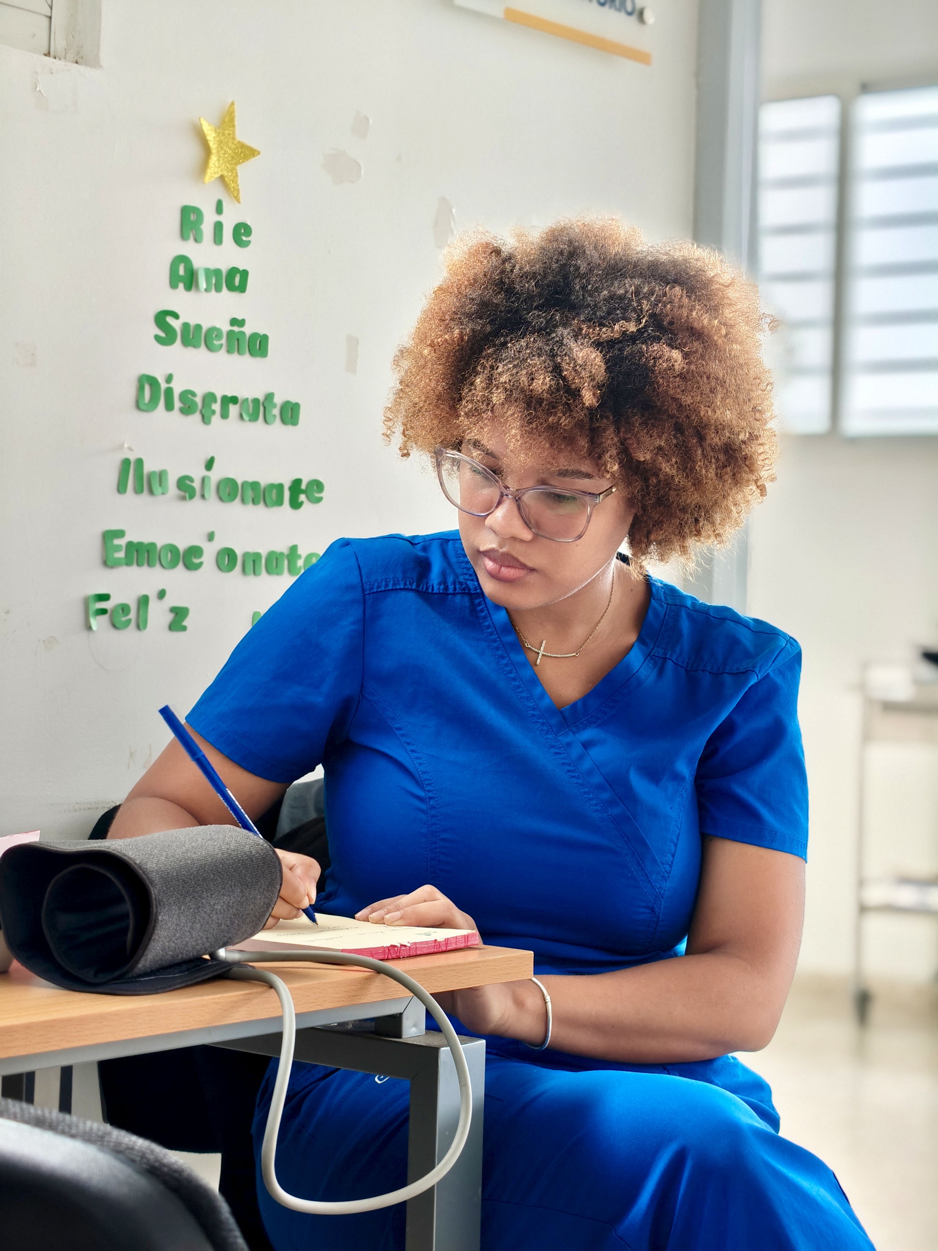 A nurse in blue scrubs writing notes by hand at a desk