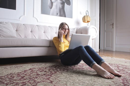 Woman in casual attire multitasking with a laptop and phone, sitting comfortably in a stylish living room.